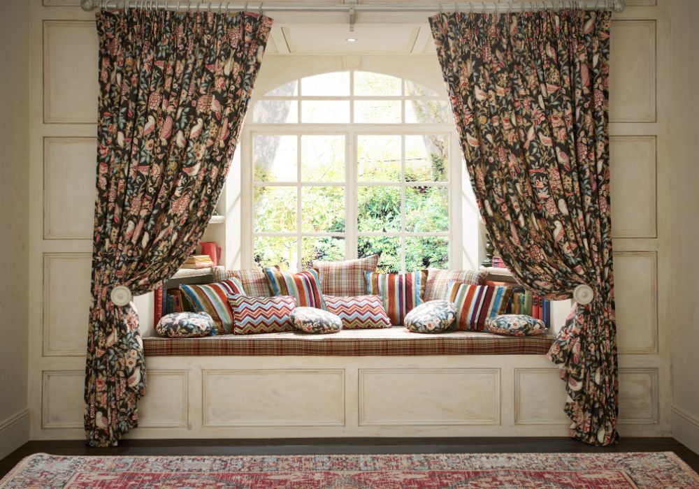 A patterned curtain framing a bay window seat with many patterned throw cushions